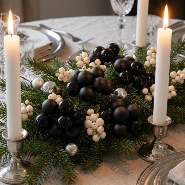 Festliche Tischdekoration mit Tannenzweigen, Weihnachtskugeln und zwei brennenden Stabkerzen in silberfarbenen Leuchtern auf einem Esstisch.