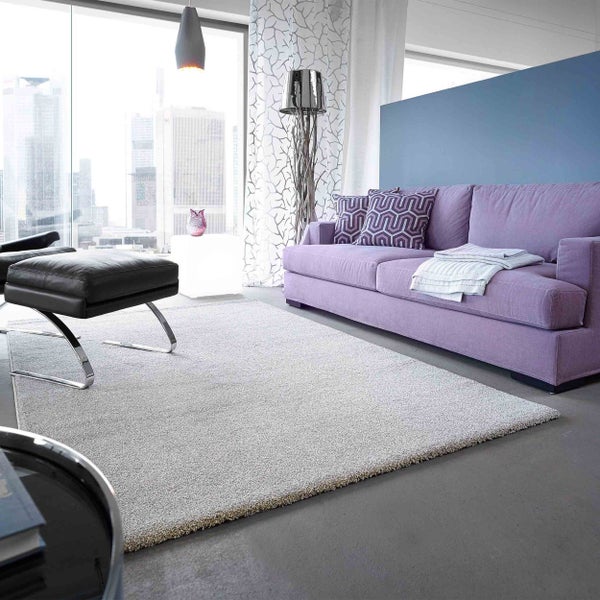 Hellgrauer Hochflor-Teppich in einem modernen Wohnzimmer mit lila Sofa, schwarzem Hocker und großer Fensterfront mit Stadtblick.