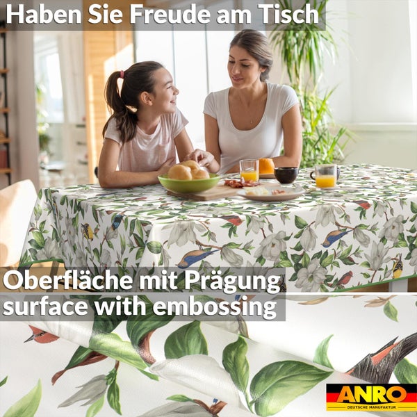 Tischdecke mit Blumen- und Vogelmuster in hellem Raum mit zwei Personen. Detailansicht der geprägten Oberfläche. ANRO Logo.