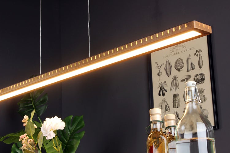 Moderne, lineare LED-Pendelleuchte aus Holz mit warmem Licht über einer Küchenszene mit Glasflaschen, Pflanzen und botanischem Wandbild.
