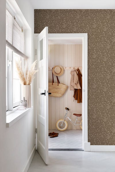 Braune Blumentapete an einer Wand in einem hellen Raum mit offener Tür zu einem Flur mit Holzpaneelen und Kinderlaufrad.