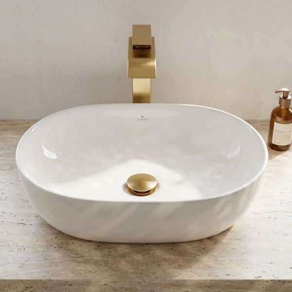 Modernes ovales Aufsatzwaschbecken aus weißer Keramik mit goldener Armatur und Ablauf auf einer beigen Waschtischplatte in Steinoptik.