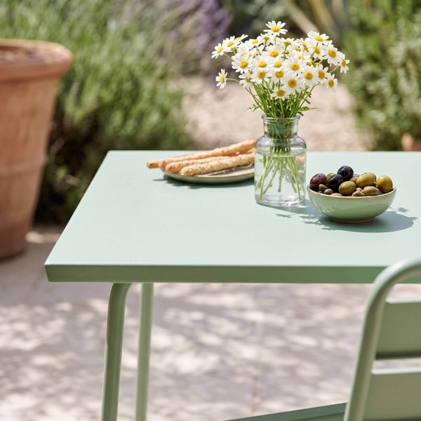 Hellgrüner Gartentisch aus Metall auf einer sonnigen Terrasse, dekoriert mit einer Blumenvase, Oliven und Brotstangen.
