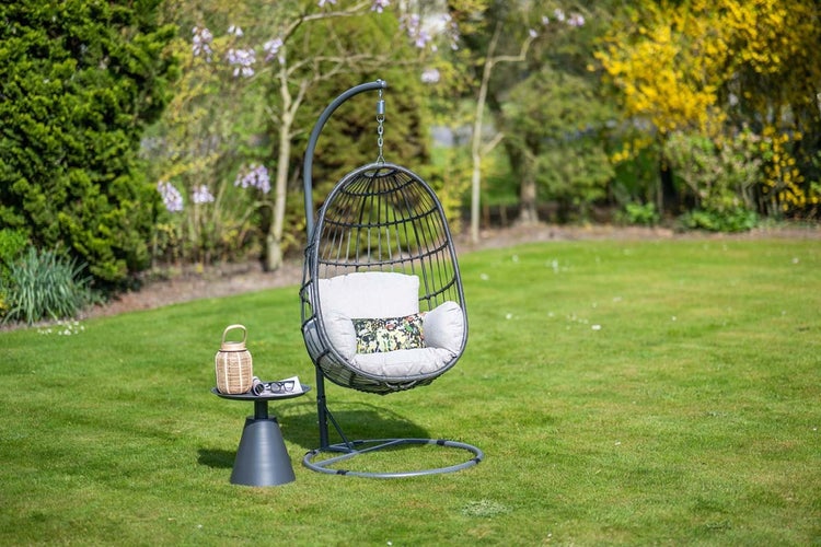 Grauer Hängesessel in Ei-Form mit Metallgestell und Kissen auf einer Rasenfläche neben einem kleinen Beistelltisch im Garten.