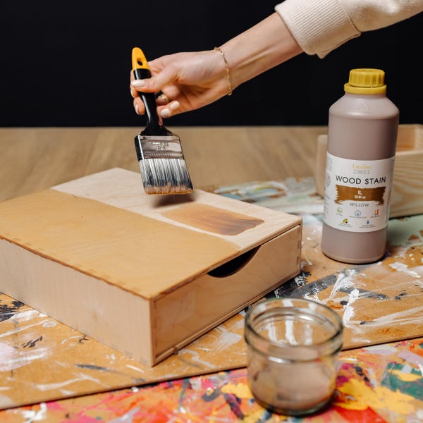 Hand trägt Holzbeize auf eine kleine Holzbox auf, neben einer Flasche Holzbeize Willow und einem Pinsel.