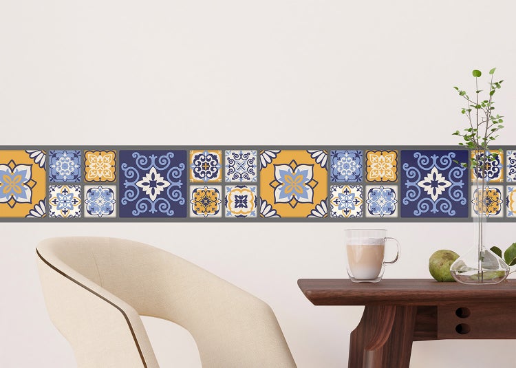 Helle Raumszene mit dekorativer Wandbordüre im mediterranen blau-gelben Fliesenmuster auf einer Wand über einem Holztisch.