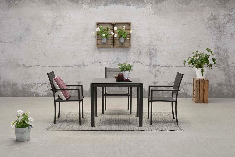 Modernes Gartenset mit quadratischem Tisch und drei Armlehnenstühlen auf einem Teppich vor einer strukturierten Wand mit Pflanzen.