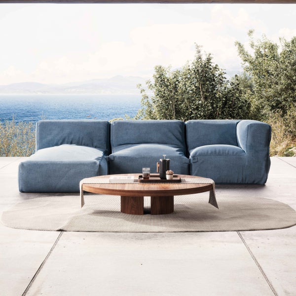 Moderne Outdoor-Lounge-Ecke mit blauem Modulsofa, rundem Couchtisch aus Holz und ovalem Teppich auf einer Terrasse mit Meerblick.