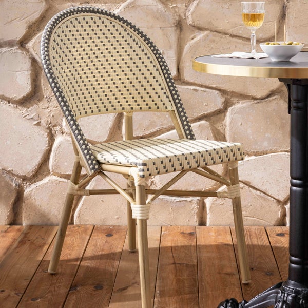 Bistrostuhl mit geflochtener Sitzfläche im Karomuster und Gestell in Bambusoptik auf einer Holzterrasse neben einem Tisch vor einer Steinwand.