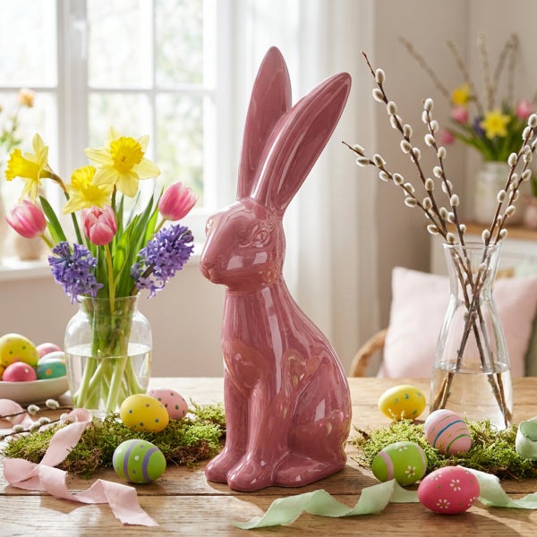 Osterdekoration mit einem sitzenden Osterhasen aus Keramik, umgeben von Frühlingsblumen, Weidenkätzchen und bemalten Ostereiern auf einem Holztisch.