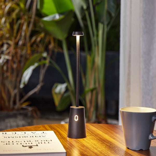 Moderne LED-Tischleuchte mit Akkubetrieb auf einem Holztisch neben einer Tasse und einem Buch, im Hintergrund Pflanzen.