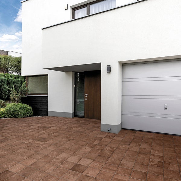 Moderne Hauseinfahrt mit braunem Pflasterstein vor einem Haus mit weißer Fassade, Eingangstür und Garagentor.
