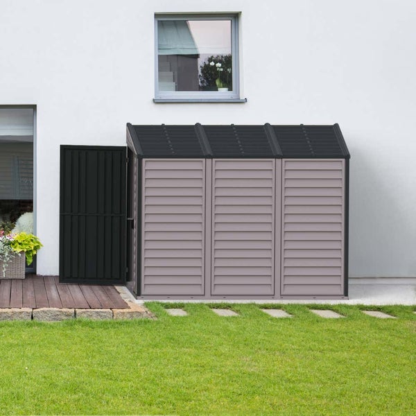 Moderne Mülltonnenbox für drei Tonnen mit Pultdach, ordentlich platziert an einer Hauswand im Garten neben einer Holzterrasse und Rasenfläche.