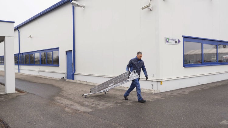 Ein Mitarbeiter in Arbeitskleidung transportiert eine Aluminiumleiter über den Parkplatz vor einem Firmengebäude.