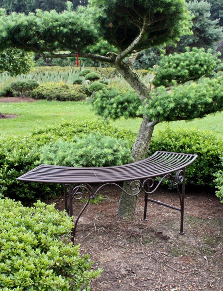 Geschwungene Gartenbank aus Metall mit verzierten Beinen in einem hellen, gepflegten Garten unter einem Nadelbaum.