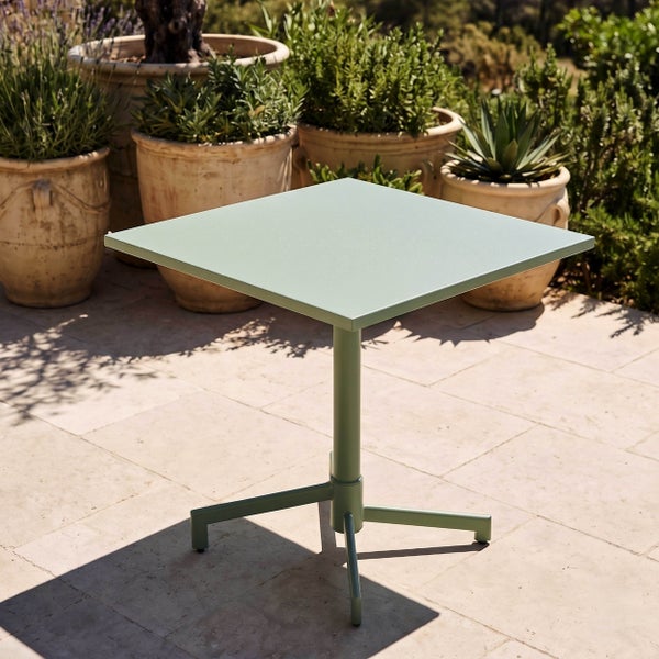 Quadratischer Bistro-Tisch aus Metall auf einer hellen Terrasse mit Pflanzen in Tontöpfen im Hintergrund.