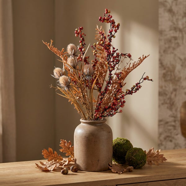 Herbstliche Blumendekoration mit Trockenblumen, Beeren und Disteln in einer Keramikvase auf einem Holztisch mit Mooskugeln und Eichenblättern.