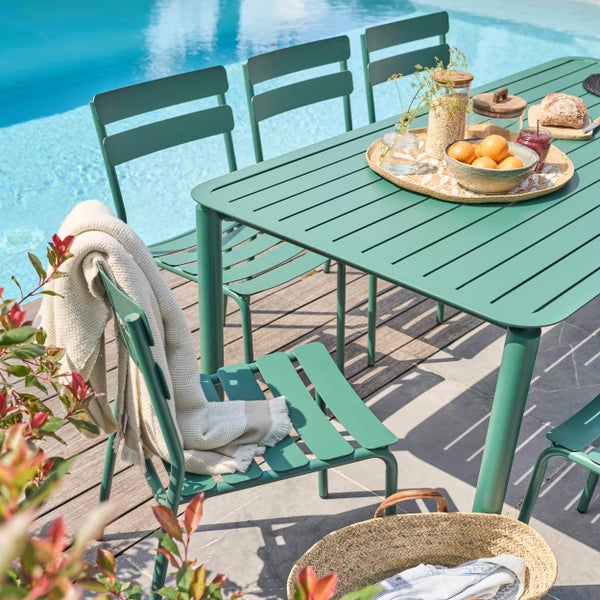 Grünes Gartenmöbel-Set aus Metall mit Tisch und Stühlen auf einer Terrasse neben einem Pool, hergerichtet für ein Frühstück.
