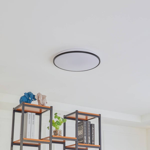 Runde, flache Deckenleuchte mit dunklem Rand in einem hellen, modern eingerichteten Raum über einem Regal mit Büchern und Dekorationsfiguren.