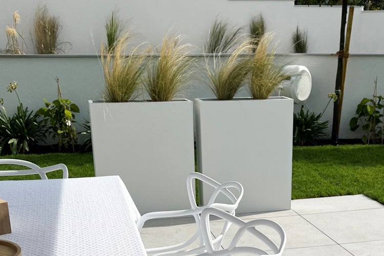Zwei hohe, rechteckige weiße Pflanzkübel mit Ziergras auf einer hellen, modernen Terrasse mit Gartenmöbeln und Rasenfläche.