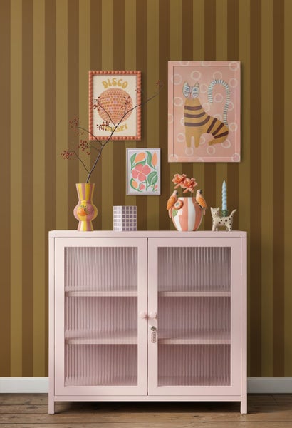 Rosafarbener Schrank mit Glastüren in Riffeloptik vor gestreifter Tapete, dekoriert mit bunten Vasen, Bildern und Wohnaccessoires.