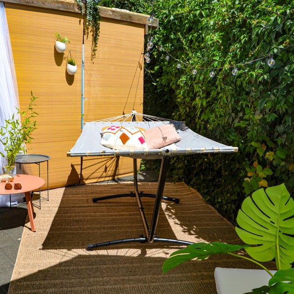 Graue Hängematte mit schwarzem Metallgestell und Kissen auf einem braunen Teppich im sonnigen Garten mit Beistelltischen und einer Hecke.