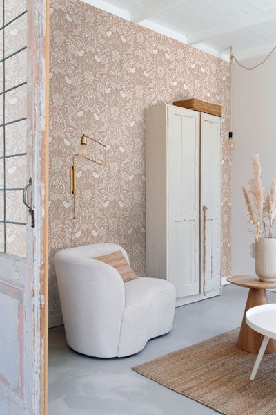 Vliestapete mit floralem Muster und Vögeln im Landhausstil, kombiniert mit einem weißen Sessel und einem rustikalen Holzschrank.