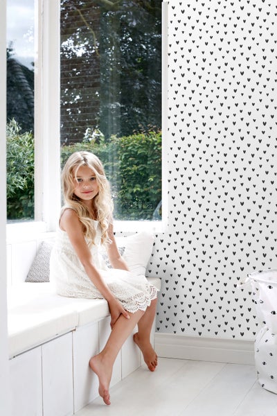 Mädchen sitzt vor einer Wand mit Herzmuster in einem hellen Raum.