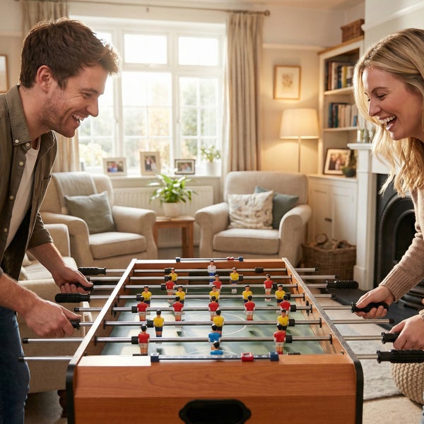 Mann und Frau spielen Tischfußball in einem hellen, organisierten Wohnzimmer mit Sesseln und Bücherregalen.