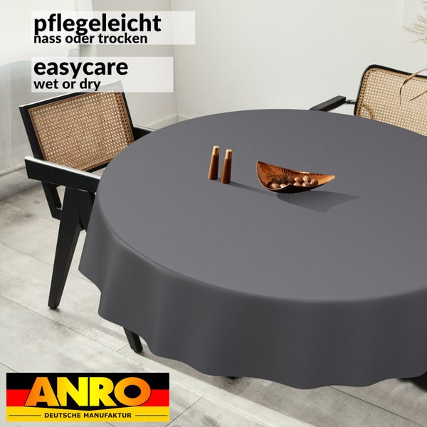 Runde, pflegeleichte Tischdecke mit Wellenrand auf einem Esstisch mit Stühlen in einem hellen Raum. ANRO Deutsche Manufaktur Logo.