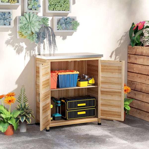 Offener Gartenschrank aus Holz mit Metall-Arbeitsplatte, gefüllt mit Werkzeugkoffern und einer Bohrmaschine, im Außenbereich neben Pflanzen.