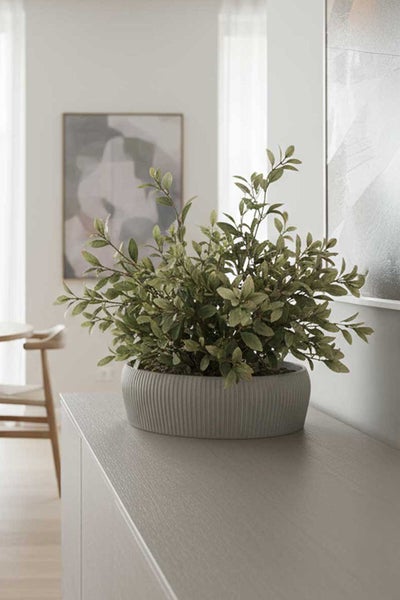 Künstliche Pflanze in grauer geriffelter Pflanzschale auf einem Sideboard in einem hellen, modernen Wohnraum mit abstrakter Wandkunst.
