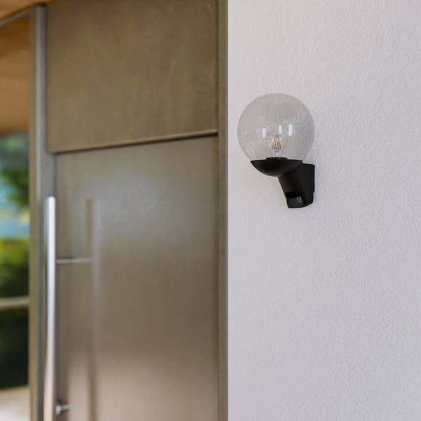 Moderne Außenwandleuchte mit kugelförmigem Glasschirm und integriertem Bewegungsmelder, montiert an einer hellen Wand neben einer Tür.