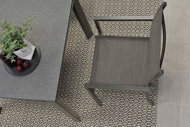 Draufsicht auf einen Gartenstuhl mit Mesh-Gewebe neben einem Gartentisch auf einem gemusterten Outdoorteppich.
