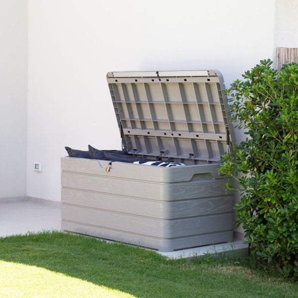 Geöffnete, graue Gartenbox mit Kissen und Decke auf Rasen vor einer Hauswand
