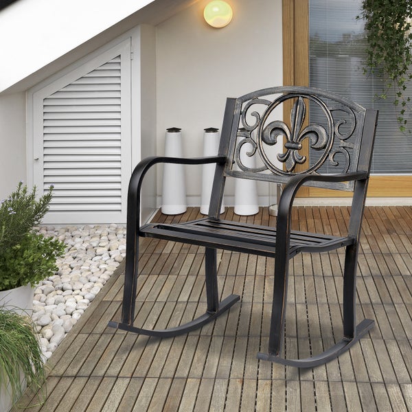 Verzierter Schaukelstuhl aus Metall mit Fleur-de-lis-Motiv auf einer hellen Terrasse mit Holzdielen, Kieselsteinen und Pflanzen.