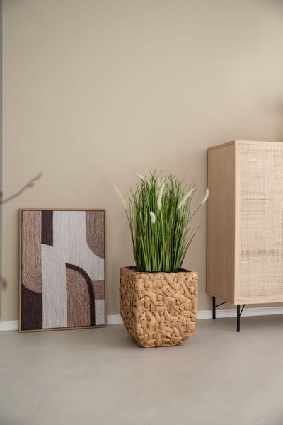 Künstliche Pflanze in quadratischem Geflecht-Übertopf, abstraktes Textilbild und Holzschrank mit Rattanfront in einem hellen Raum.