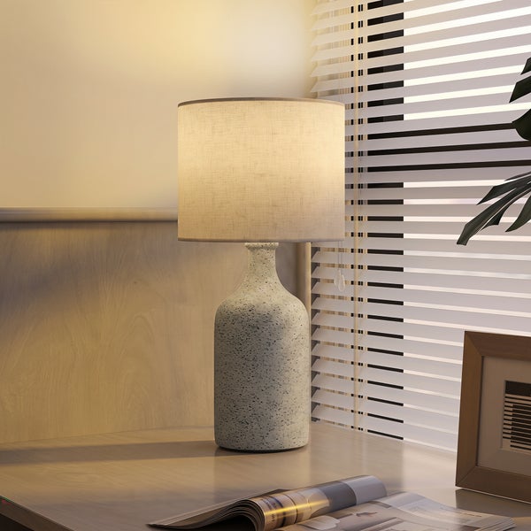 Tischleuchte mit zylindrischem Stoffschirm und gemustertem Keramikfuß auf einem Holztisch neben einer Pflanze in einem Topf.