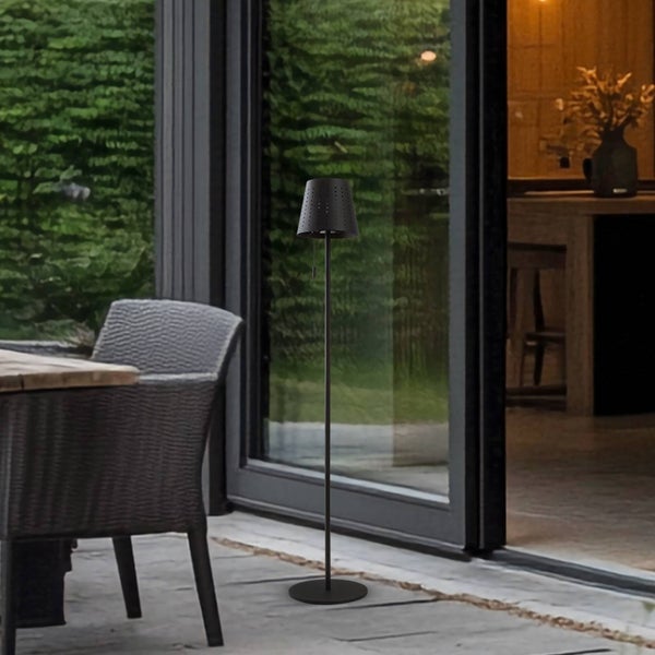 Moderne schwarze Außenstehlampe mit perforiertem Schirm auf einer Steinterrasse neben einer Glastür und Gartenmöbeln.