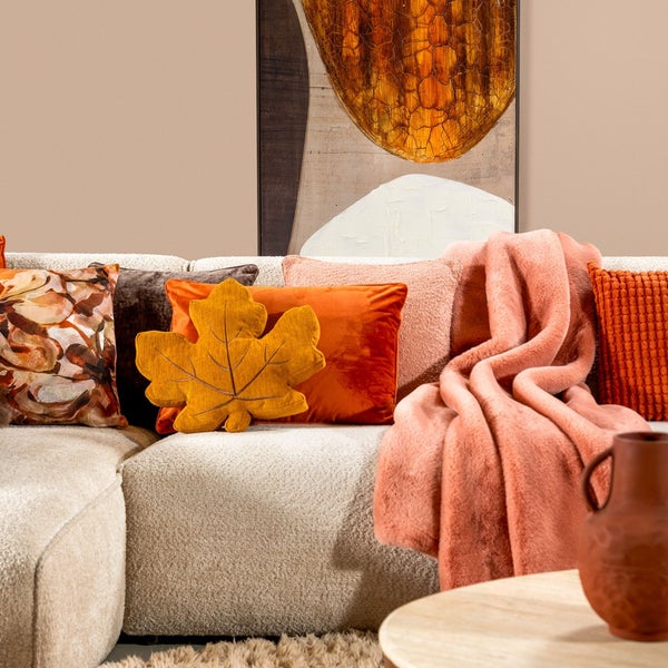 Gemütliches Sofa mit verschiedenen Zierkissen in herbstlichen Farben, einer Kuscheldecke und einem Kissen in Blattform vor einem Wandbild.