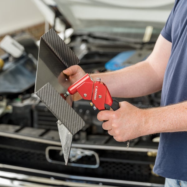 Person nutzt rote Arrow Handnietzange an Karbonbauteil vor einem PKW-Motorraum.