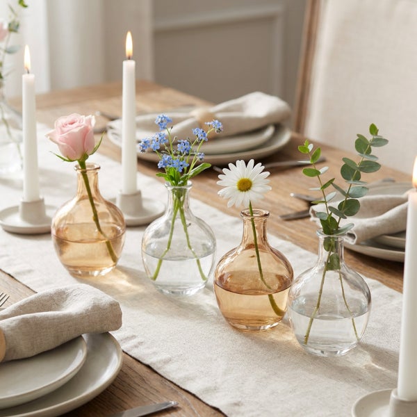Set aus vier kleinen Glasvasen in verschiedenen Formen auf einem gedeckten Holztisch mit Blumen und Kerzen.
