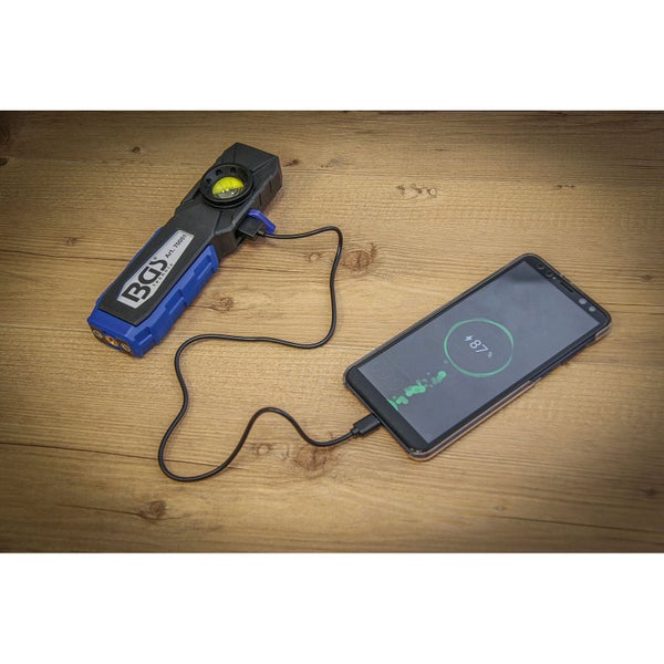 BGS Technic LED-Arbeitsleuchte mit Powerbank-Funktion lädt ein Smartphone über ein USB-Kabel auf einer Holzoberfläche.