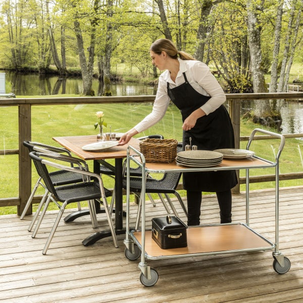 Szene im Freien mit einer Kellnerin, die einen Tisch deckt, mit Servierwagen, Stühlen und Tisch auf einer Terrasse mit Blick auf einen See