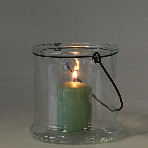 Brennende grüne Kerze in einem zylindrischen Windlicht aus Glas mit schwarzem Metallbügel.
