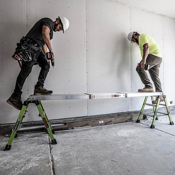 Zwei Handwerker nutzen ein Gerüstsystem aus zwei Trittleitern und einer Arbeitsplattform zur Arbeit an einer Trockenbauwand.