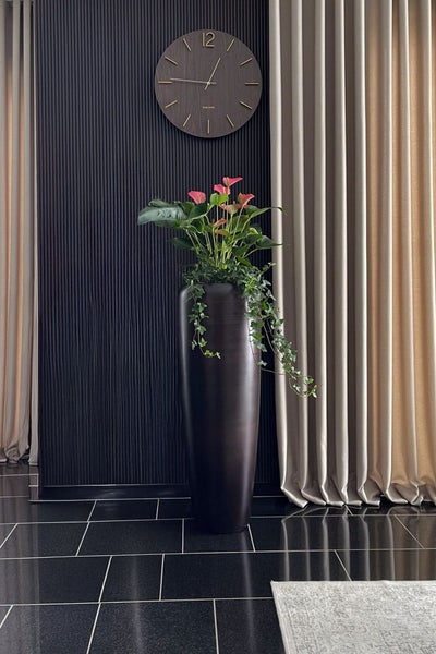 Hohe dunkle Bodenvase mit Anthurie und Efeu vor einer dunklen Lamellenwand mit runder Holzuhr und Vorhängen.