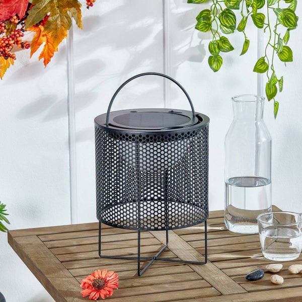 Schwarze Solar-Laterne aus Metall mit Gitterstruktur auf einem Holztisch neben einer Glaskaraffe und herbstlicher Dekoration.