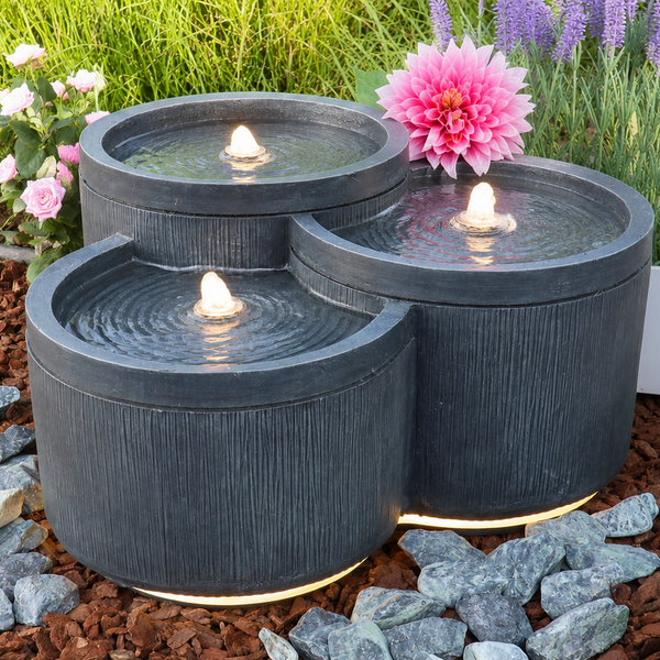 Dreistufiger Kaskadenbrunnen mit strukturierter Oberfläche und LED-Beleuchtung in einem Gartenbeet mit Mulch und Blumen.