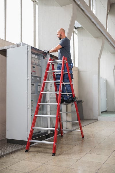 Handwerker steht auf einer stabilen Stehleiter und arbeitet an einem großen Schaltschrank in einer industriellen Umgebung.